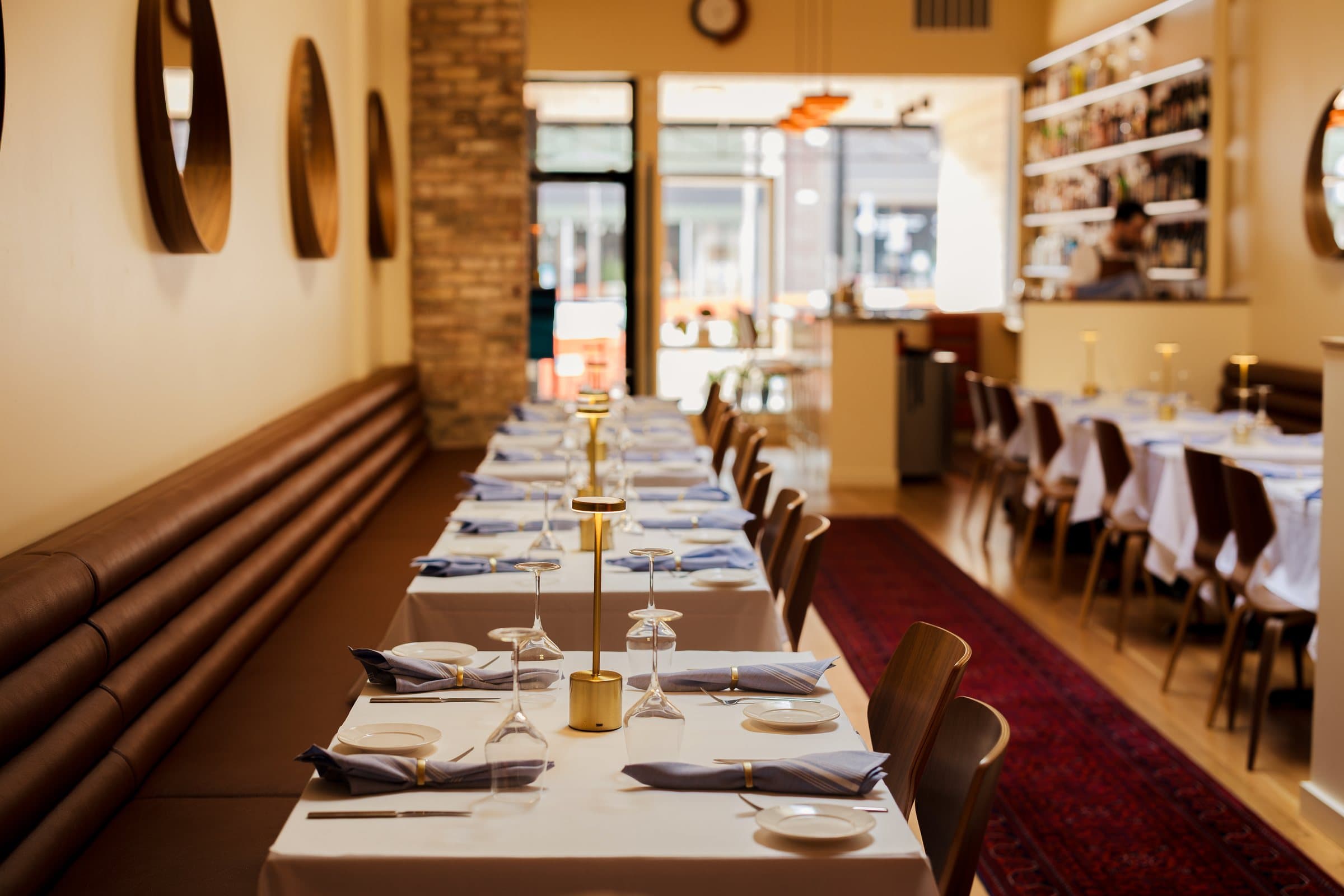 The long dining room at Kismet, with a leather banquette, round walnut mirrors and brass lamps on white tablecloths